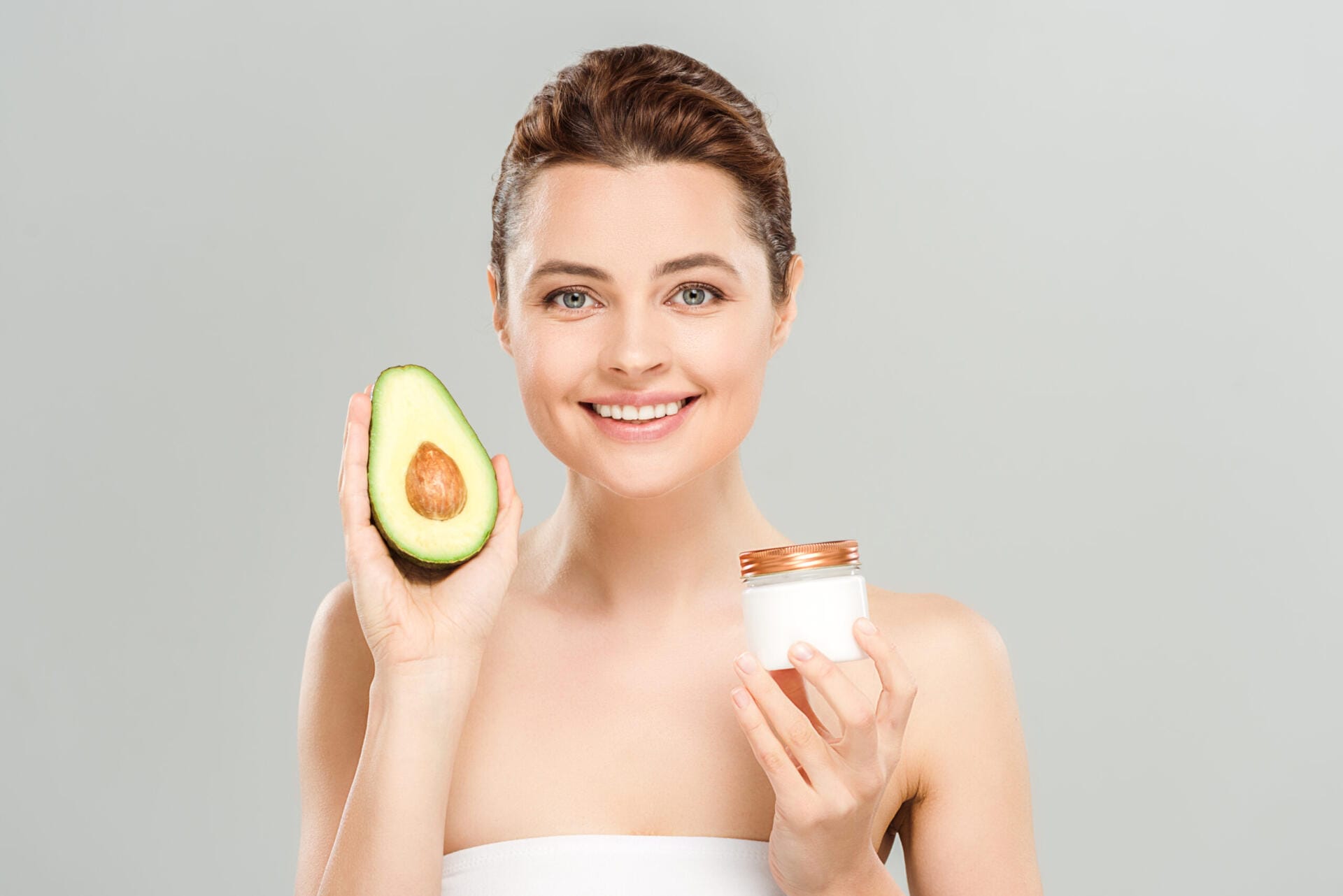 cheerful woman holding half of avocado and container with cosmetic cream isolated on grey